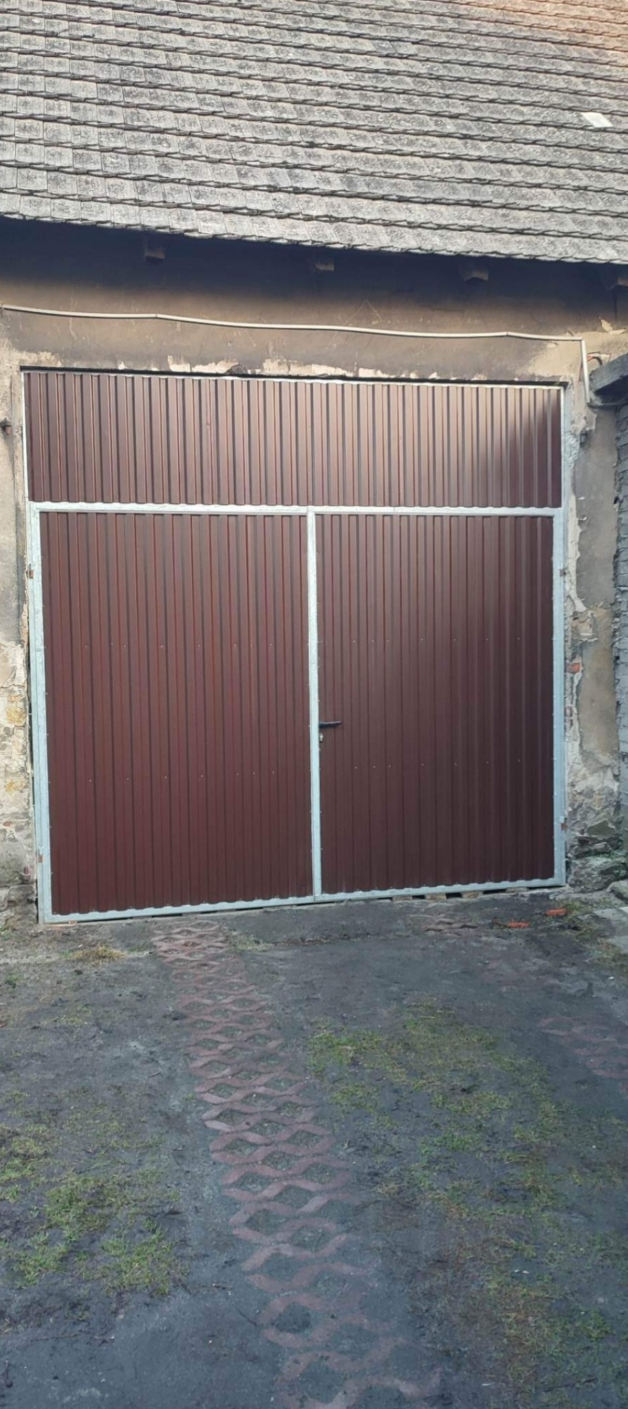 Brązowa, dwuskrzydłowa brama garażowa z metalowych paneli, z jasną ramą, zamontowana w starej, częściowo otynkowanej ścianie budynku z dachówką. Na dole widoczny podjazd z ażurowych płyt.