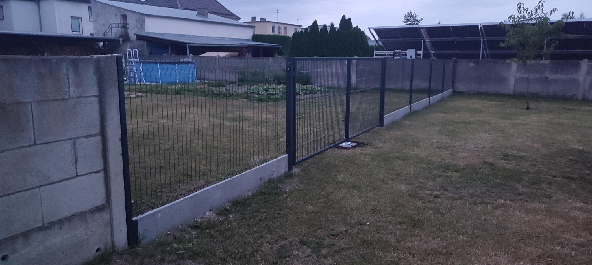 Nowoczesne ogrodzenie panelowe z podmurówką betonową, widok z perspektywy trawnika, w tle dom z basenem i panele słoneczne.