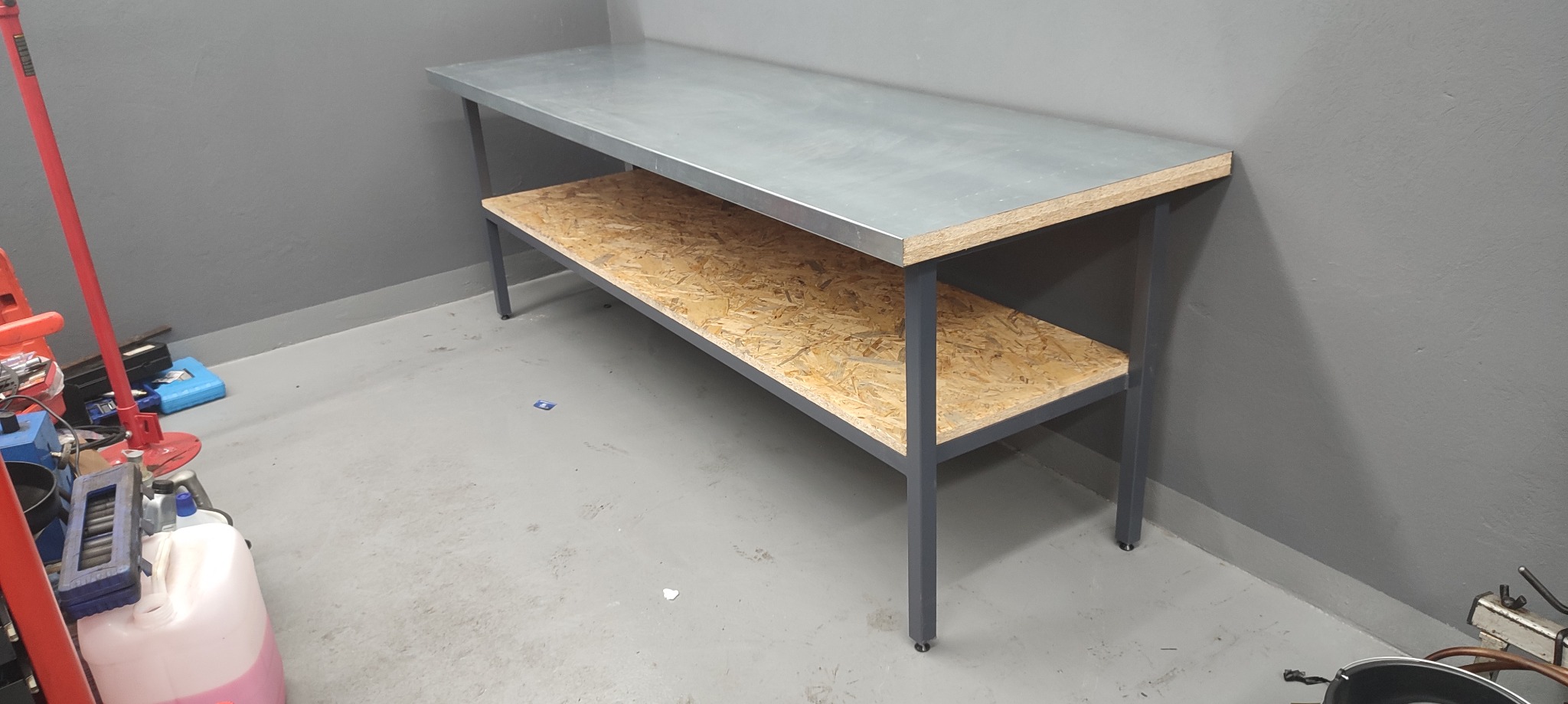 Metalowy stół warsztatowy z blatem ze stali nierdzewnej i półką z płyty OSB, ustawiony w rogu szarego garażu, obok narzędzi i pojemników.