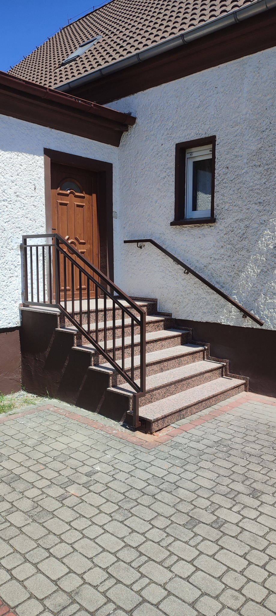 Zewnętrzne schody wejściowe z brązowego granitu z metalową balustradą w kolorze brązowym, prowadzące do drewnianych drzwi w białym budynku z oknem.