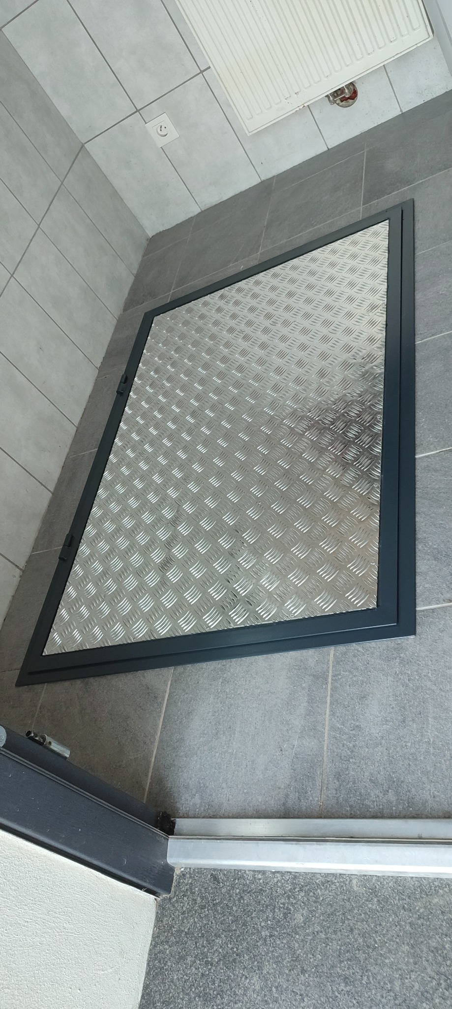 Metalowa klapa rewizyjna z blachy ryflowanej, wbudowana w podłogę z szarych płytek, widoczna z góry pod kątem.