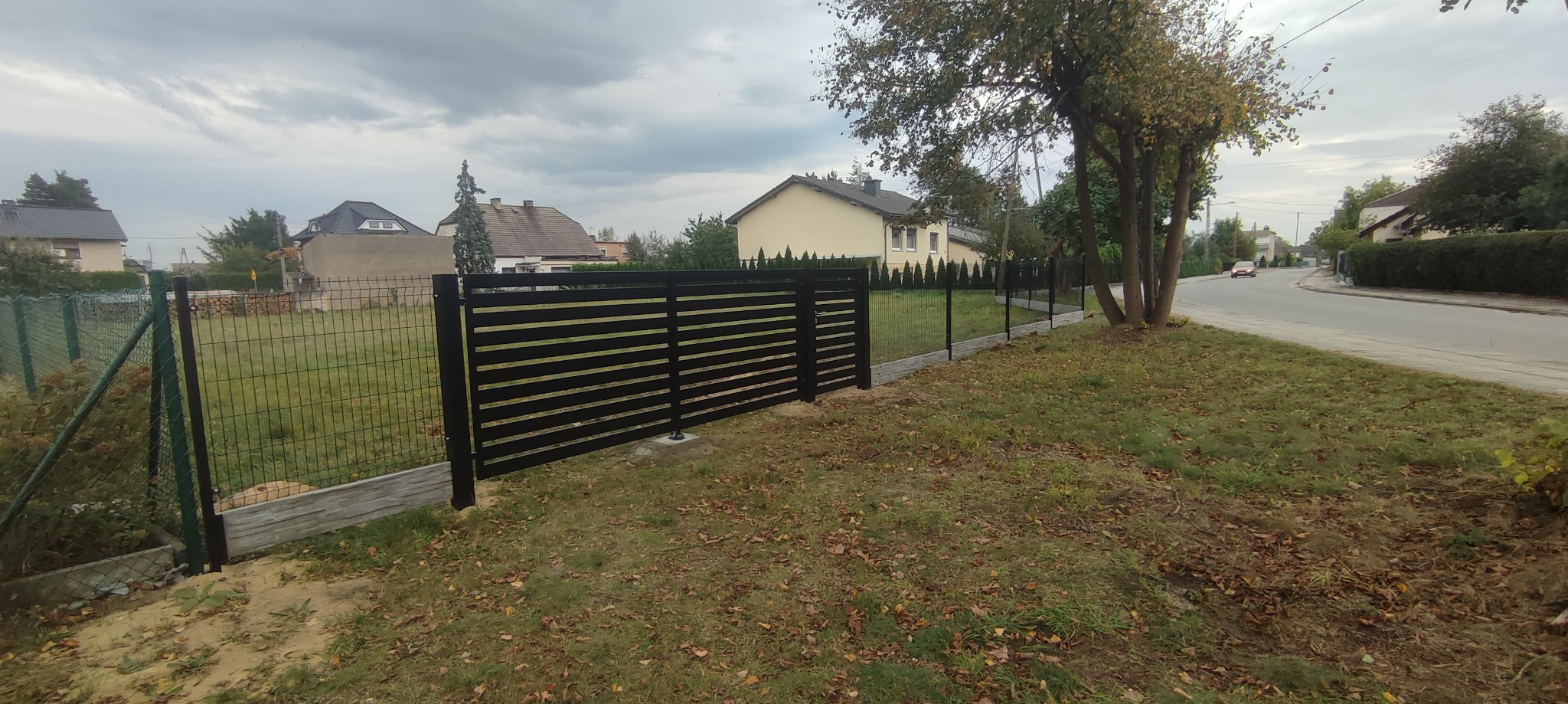 Nowoczesne, czarne ogrodzenie z poziomych paneli na betonowej podmurówce, połączone z siatką ogrodzeniową, na tle domów jednorodzinnych i pochmurnego nieba.