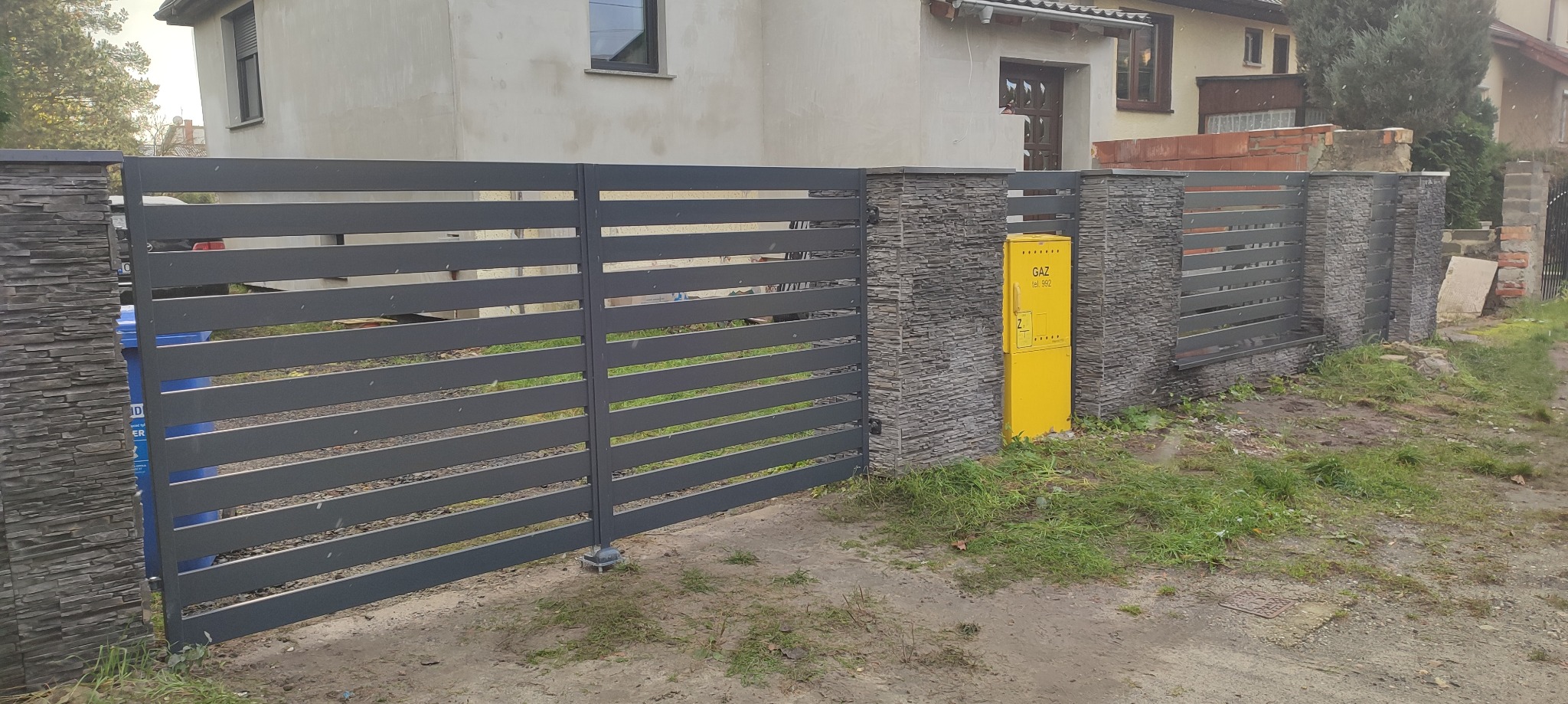 Nowoczesne ogrodzenie panelowe w Opolu z szarymi, poziomymi przęsłami i kamiennymi słupkami, z widoczną skrzynką gazową w jaskrawym kolorze.