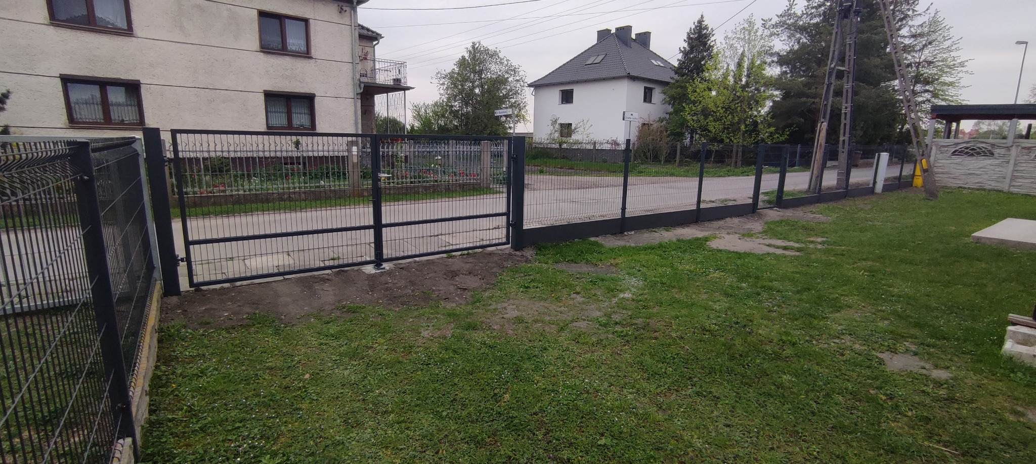 Nowoczesne ogrodzenie panelowe z otwartą bramą wjazdową, widok na ulicę i domy w tle.