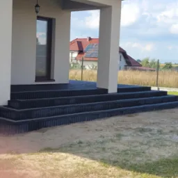 Schody wejściowe do domu wyłożone ciemną kostką brukową, widoczne z poziomu gruntu, z piaskiem i trawą w tle, w oddali dom z czerwoną dachówką i panelami słonecznymi.