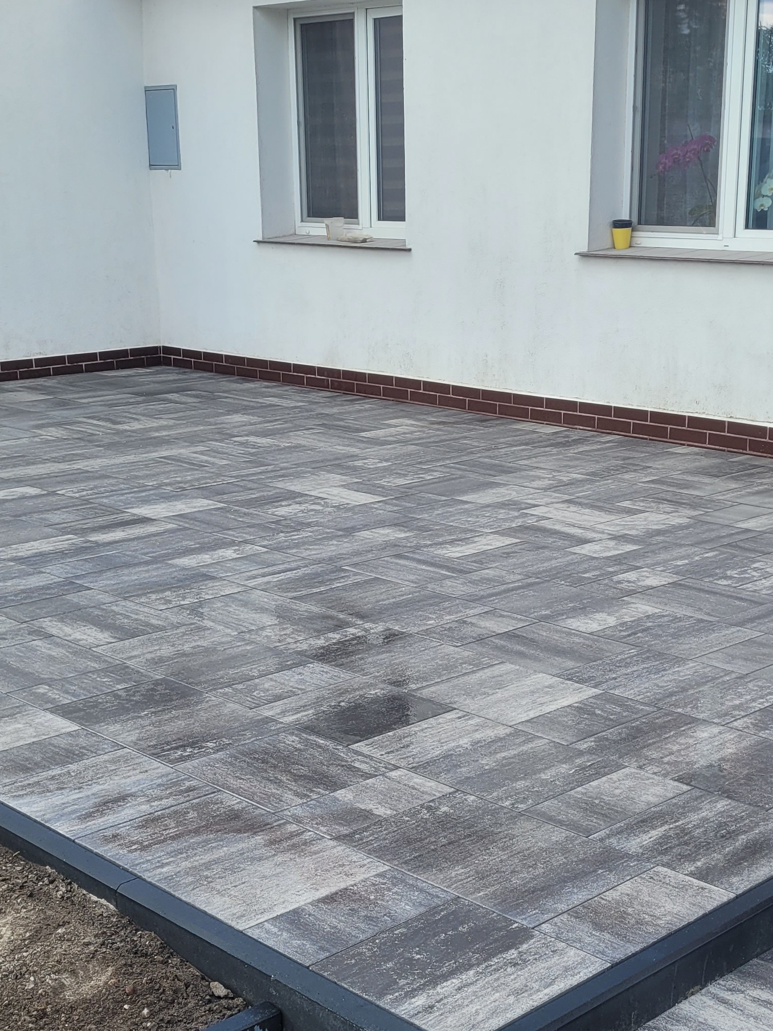 Szara, ułożona kostka na posesji, widok z góry, obrzeża z czarnym krawężnikiem, fragment białej ściany budynku z dwoma oknami w tle.