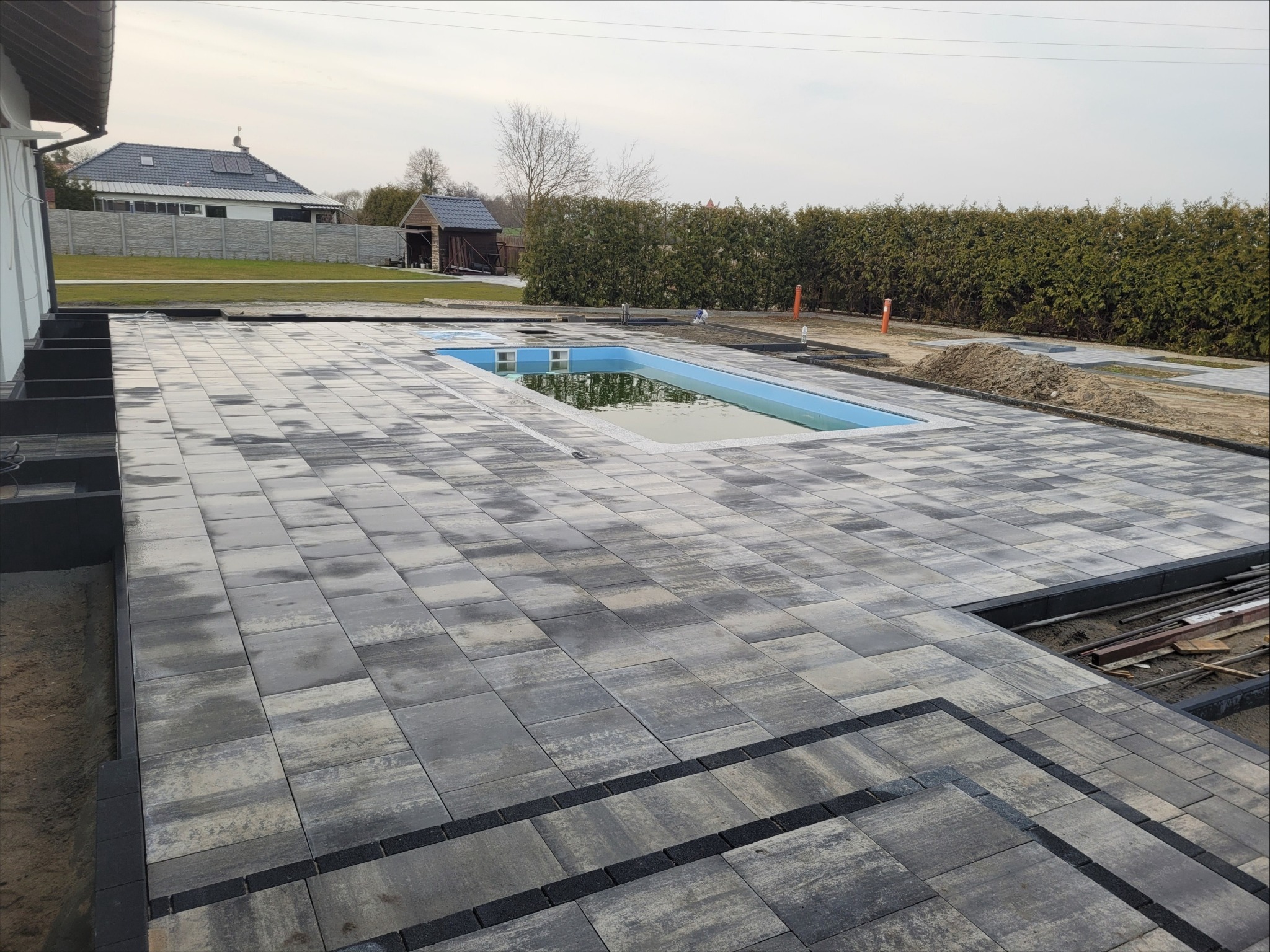 Szare kostki brukowe ułożone wokół basenu, widoczne niedokończone fragmenty, w tle dom jednorodzinny i zielony żywopłot.