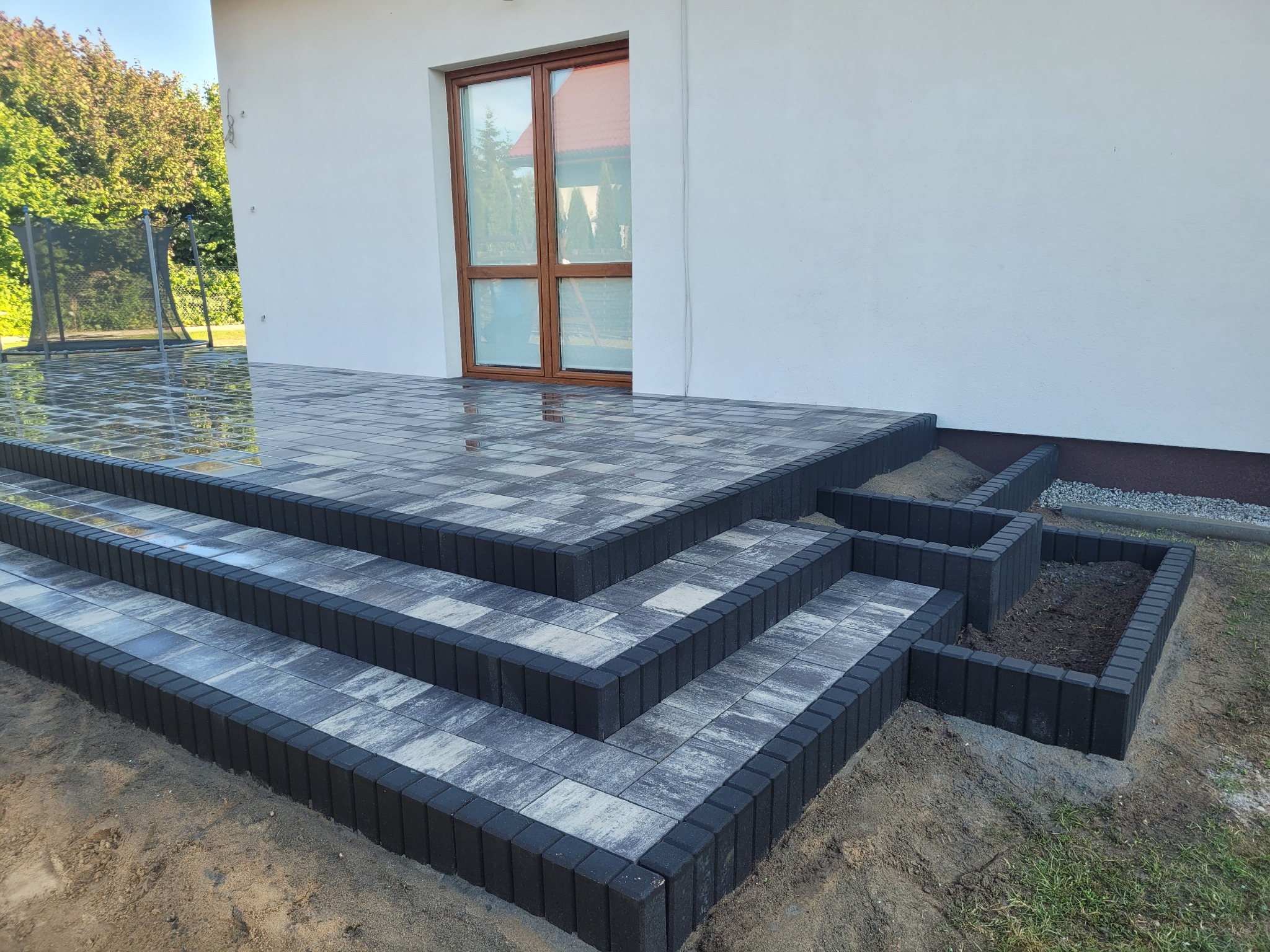 Schody wejściowe do domu wyłożone szarą kostką brukową z ciemnoszarą obwódką, obok rabata kwiatowa z obrzeżem z tej samej kostki, widoczne odbicia w mokrej nawierzchni.