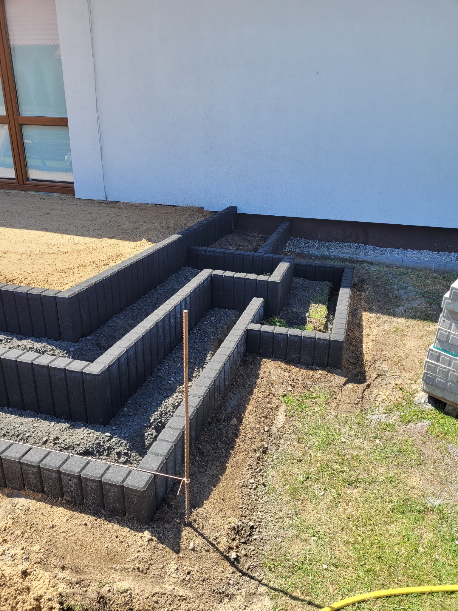 Formowanie rabat kwiatowych z szarej kostki obrzeżowej, widoczne etapy prac przygotowawczych, częściowo wypełnione ziemią i gruzem, w tle biała ściana budynku i paleta z kostką.