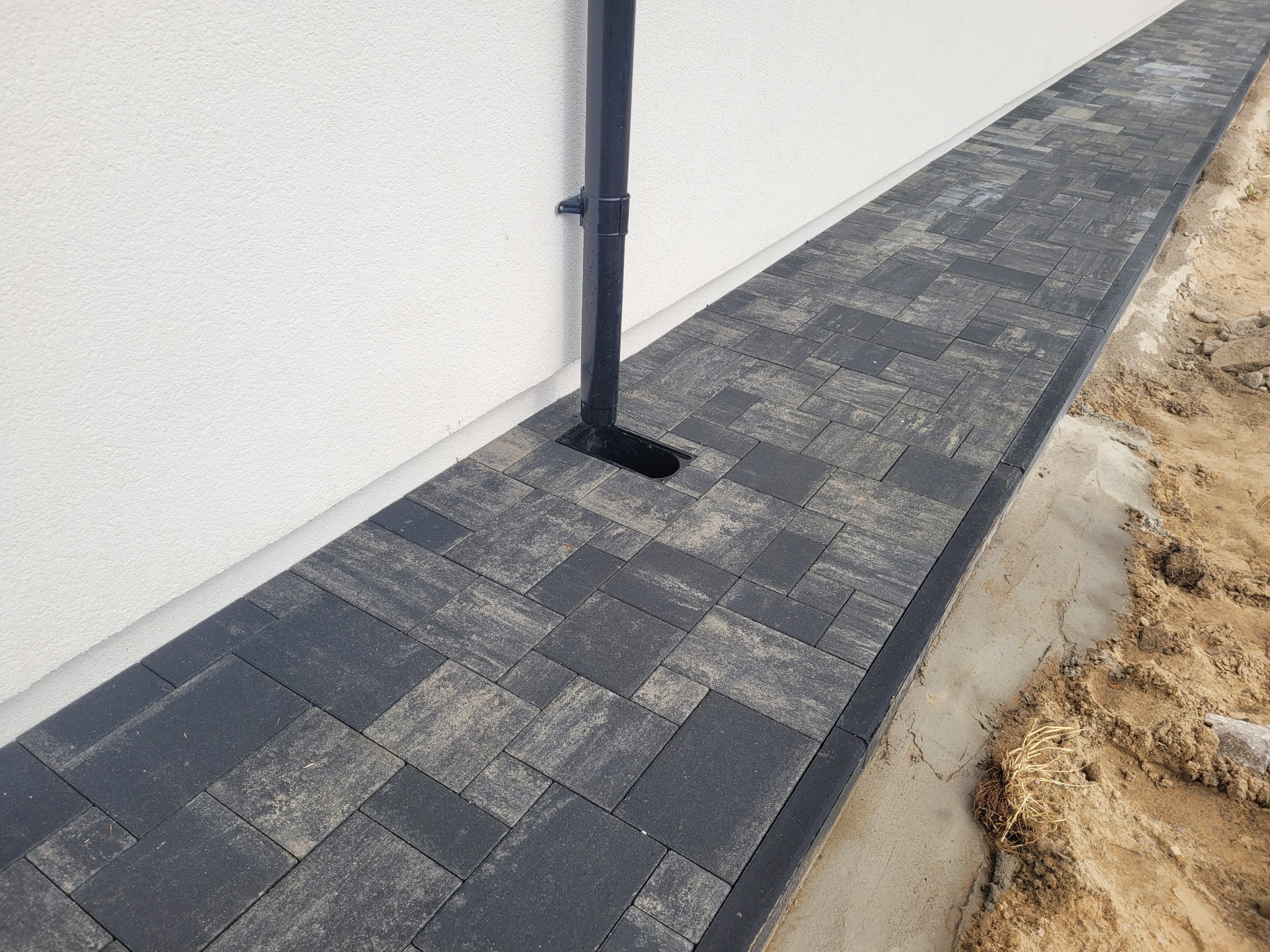Ścieżka z ciemnoszarej kostki ułożonej przy białej ścianie budynku, z rurą spustową odprowadzającą wodę deszczową, widoczne ślady piasku na kostce.