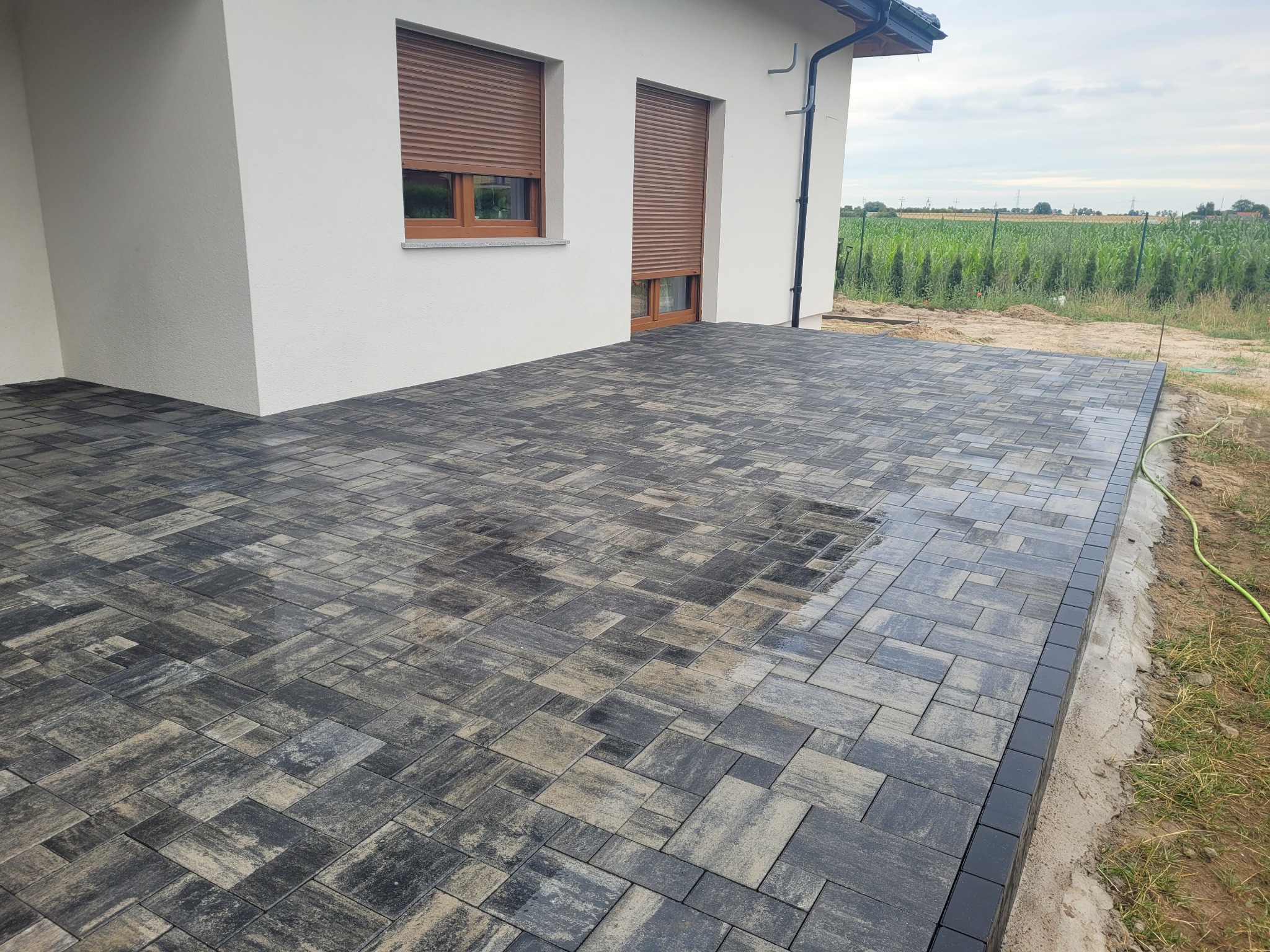 Nowo ułożony taras z szaro-beżowej kostki, obrzeża z ciemnych kostek, widok na biały budynek z brązowymi roletami i zielone pole w tle.