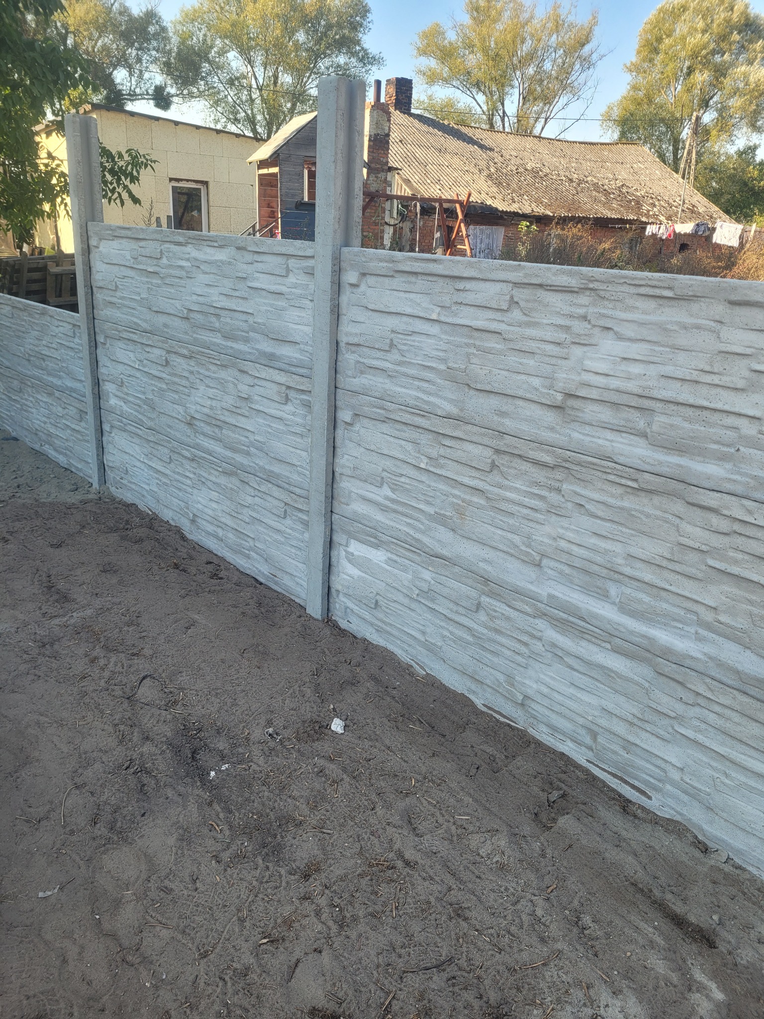 Nowo postawiony, prefabrykowany płot betonowy o teksturze kamienia, widok z bliska na ziemię przy fundamencie, w tle dom z kominem i drabina oparta o ścianę.