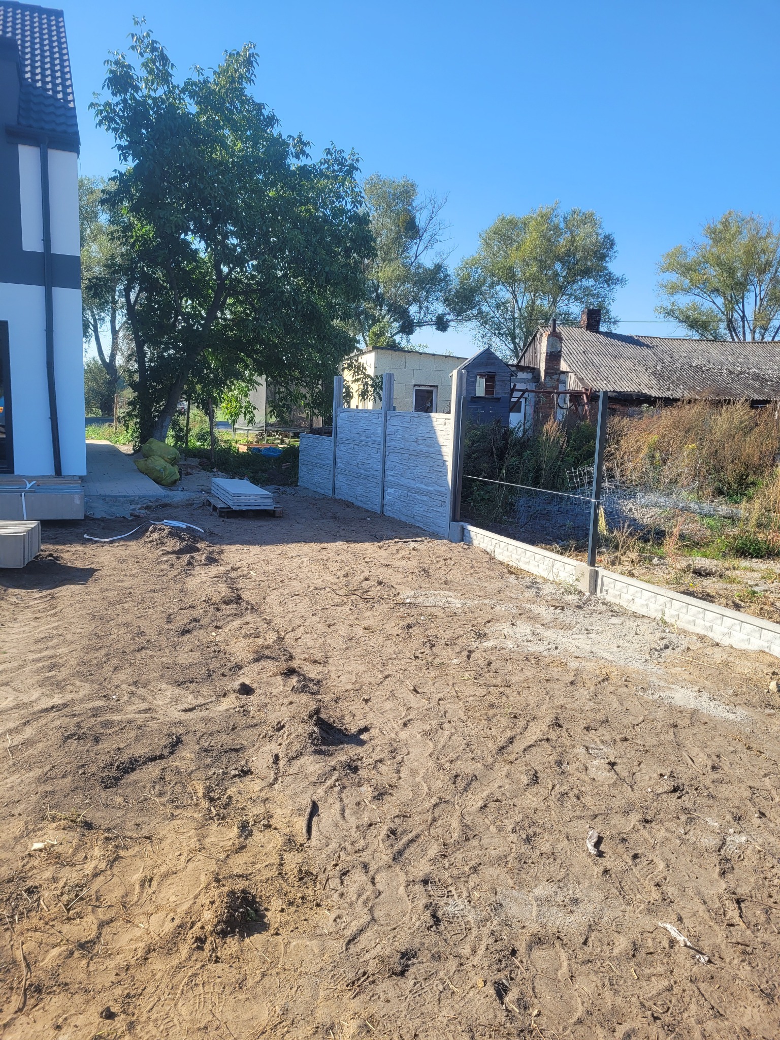 Wznoszenie betonowego ogrodzenia panelowego na nierównym terenie, widoczne przygotowane płyty i słupki, w tle nowy dom i zabudowania gospodarcze.