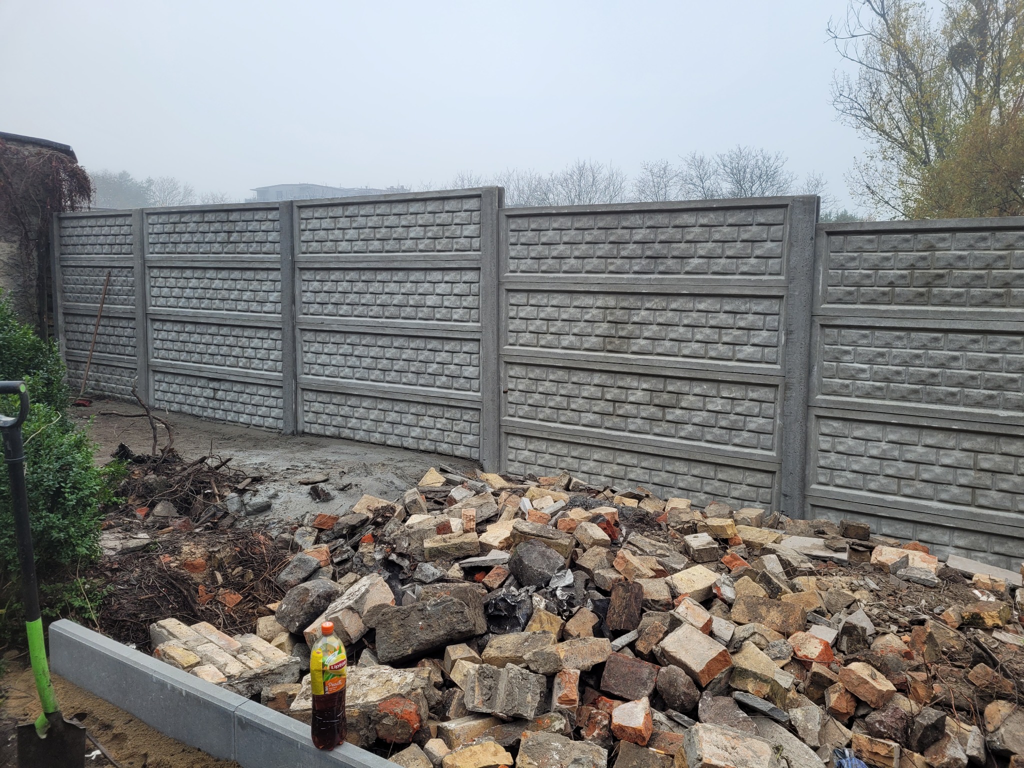 Montaż prefabrykowanego betonowego ogrodzenia panelowego z widocznym stosem gruzu i butelką napoju w tle zamglonego krajobrazu.