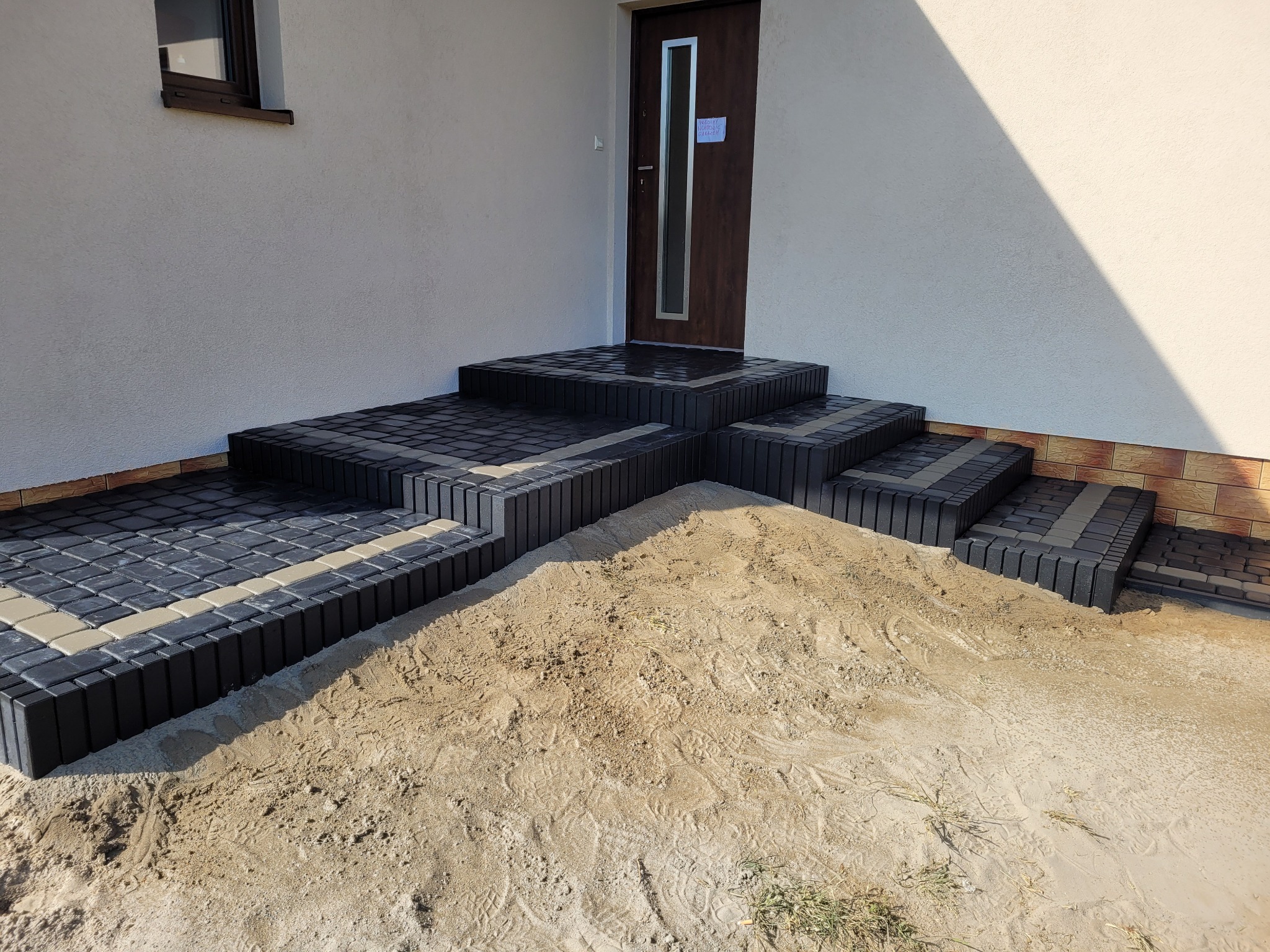 Schody wejściowe do domu w Mirakowie wyłożone czarno-beżową kostką brukową, częściowo zasypane piaskiem.