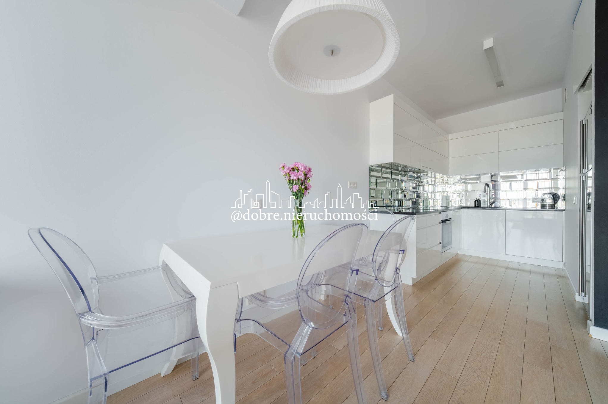 Jasne wnętrze: biały stół z kwiatami i przezroczystymi krzesłami, w tle błyszcząca kuchnia z lustrzanymi płytkami. Minimalistyczny design, drewniana podłoga.