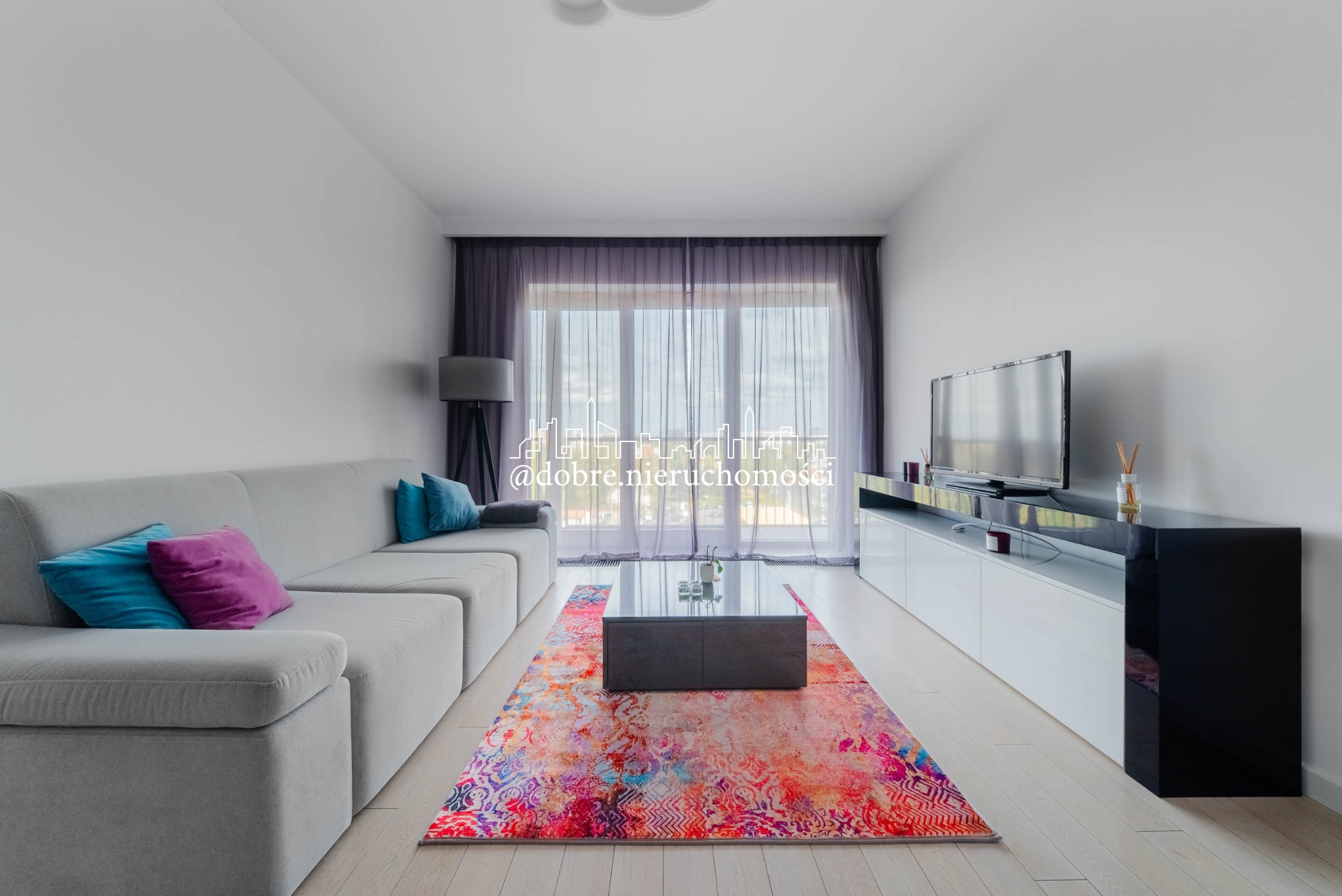 Jasne wnętrze salonu z szarą sofą, kolorowym dywanem i czarno-białą szafką RTV. Widok na okno z firanami. Nowoczesny design z akcentami kolorystycznymi.