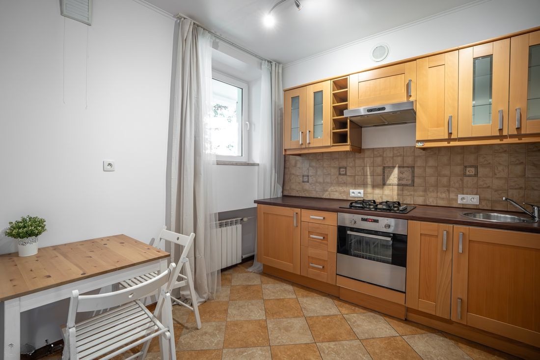 Jasna kuchnia z drewnianymi szafkami, brązowym blatem i stołem z białymi krzesłami. Okno z firanką. Płytki podłogowe w szachownicę. Ujęcie z perspektywy.