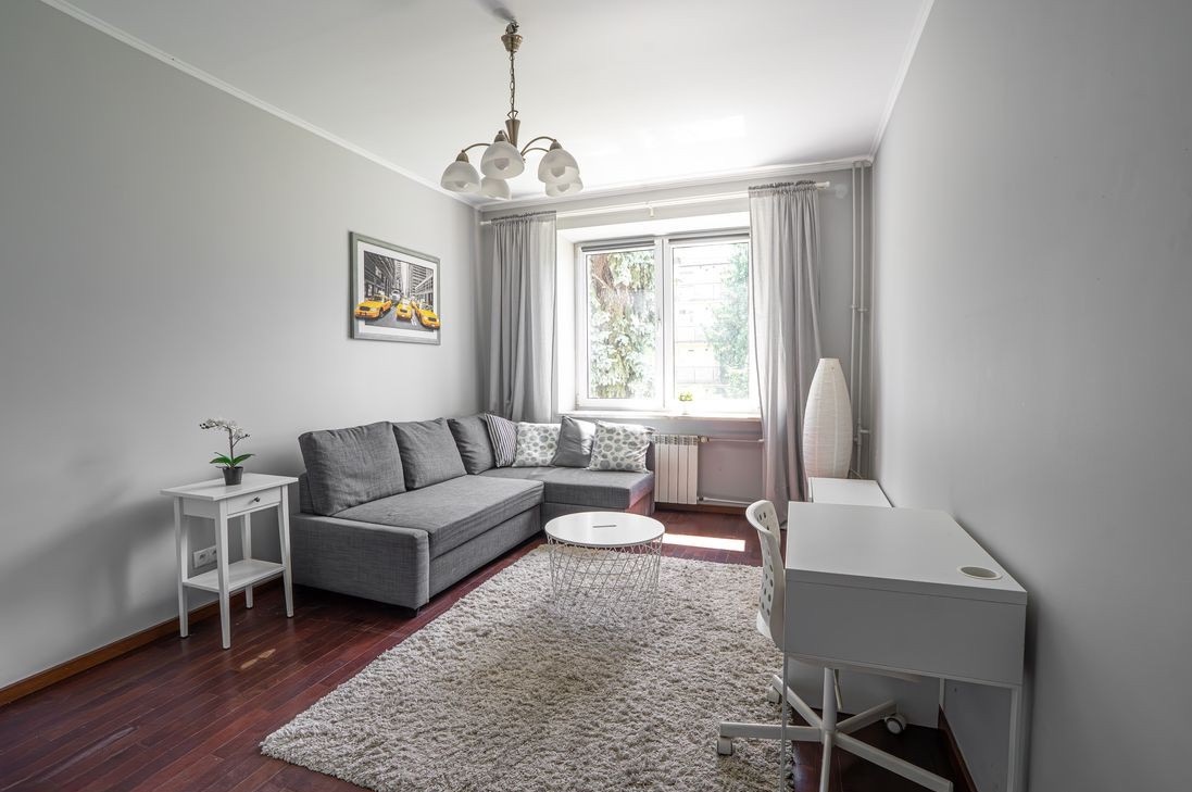 Jasne, nowocześnie urządzone wnętrze pokoju z szarą sofą narożną, białym dywanem i biurkiem. Na ścianie wisi obraz z żółtymi taksówkami.