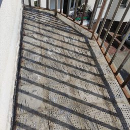 WoodyMAXX Sergiusz Proń - Balkon w trakcie remontu, widoczna stara, zniszczona powierzchnia z pozostałościami po poprzedniej warstwie, metalowa balustrada rzuca cienie na podłoże.