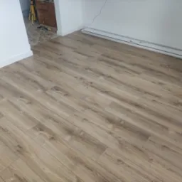 Ukończona podłoga z paneli laminowanych w odcieniach brązu, ułożona w jasnym pomieszczeniu z białymi ścianami i listwami przypodłogowymi. Widoczny fragment grzejnika przy ścianie.