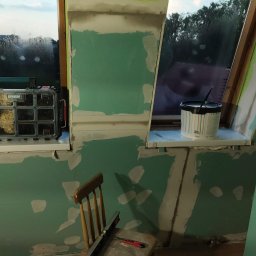 WoodyMAXX Sergiusz Proń - Poddasze w trakcie remontu: zielone ściany z gipsem, okna dachowe z widokiem na zewnątrz, organizer Erbauer z drobnymi elementami na parapecie, wiadro z farbą, poziomica i nóż na krześle.