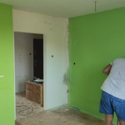 WoodyMAXX Sergiusz Proń - Mężczyzna w czerwonej czapce maluje ścianę na jaskrawy zielony kolor w pokoju w trakcie remontu; widoczne gniazdka elektryczne, kable i fragmenty ściany w innym kolorze.