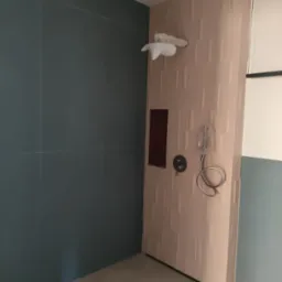 Nowoczesna kabina prysznicowa z beżową, teksturowaną ścianą, zamontowaną armaturą i niebiesko-szarymi płytkami na ścianie bocznej, zabezpieczona folią.