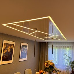 WoodyMAXX - Nowoczesny sufit napinany z podświetleniem LED w geometryczne wzory, odbijający elementy wystroju wnętrza. Widoczne wiszące lampy i dekoracje.