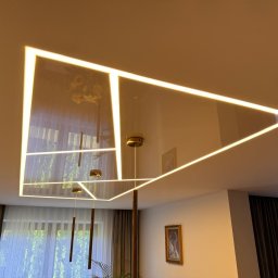 WoodyMAXX - Błyszczący sufit napinany z oświetleniem LED w kształcie geometrycznym, odbijający okno i wiszące lampy. Nowoczesny design wnętrza w ciepłej tonacji.