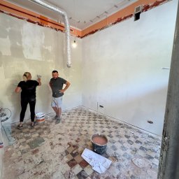 WoodyMAXX Sergiusz Proń - Prace wykończeniowe w pomieszczeniu z zachowaną, starą posadzką w szachownicę. Kobieta nakłada gładź na ścianę, obok stoi mężczyzna. Widoczne wiadro z gładzią.