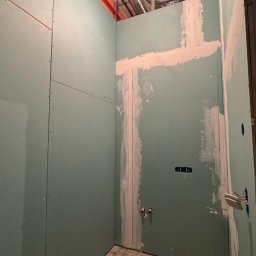 WoodyMAXX Sergiusz Proń - Wnętrze w trakcie prac: ściany z płyt gipsowo-kartonowych, widoczne instalacje, częściowo zagruntowane spoiny, rury wentylacyjne pod sufitem. Faza przygotowawcza przed wykończeniem.