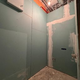 WoodyMAXX Sergiusz Proń - Wnętrze w trakcie montażu płyt karton-gips, widoczne instalacje, częściowo wyszpachlowane łączenia. Na podłodze resztki materiałów budowlanych.