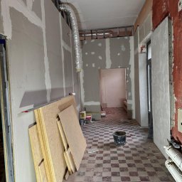 WoodyMAXX Sergiusz Proń - Wnętrze w trakcie remontu: widoczne płyty karton-gips na ścianach, metalowe profile, rury wentylacyjne, płyty OSB i podłoga w kratkę. Prace w toku.