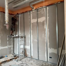 WoodyMAXX Sergiusz Proń - Montaż ścianki działowej z płyt gipsowo-kartonowych na metalowym stelażu. Widoczny pracownik na rusztowaniu i instalacja wentylacyjna pod sufitem.