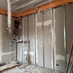 WoodyMAXX Sergiusz Proń - Montaż płyt karton-gips na metalowym stelażu. Pracownik na rusztowaniu, widoczne przewody wentylacyjne pod sufitem. Prace wykończeniowe w trakcie realizacji.