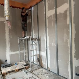 WoodyMAXX - Montaż ścianki działowej z profili i płyt gipsowych. Mężczyzna na rusztowaniu przykręca płytę. Widoczne narzędzia i materiały budowlane. Pomieszczenie w trakcie prac wykończeniowych.
