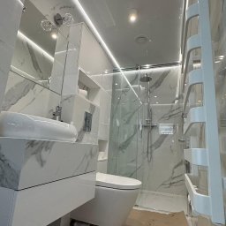 WoodyMAXX Sergiusz Proń - Nowoczesna łazienka w bieli i marmurze: podświetlane lustro, szklana kabina prysznicowa, wisząca toaleta, grzejnik drabinkowy. Podłoga zabezpieczona kartonem.