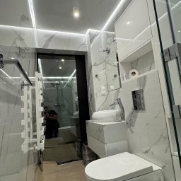 WoodyMAXX Sergiusz Proń - Nowoczesna łazienka w bieli i marmurze z podświetleniem LED, widok na kabinę prysznicową, odbicie osoby robiącej zdjęcie w lustrze. Minimalizm i elegancja.