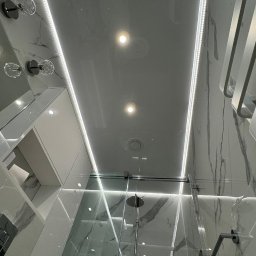 WoodyMAXX - Nowoczesna łazienka z podświetlanym sufitem napinanym, marmurowe płytki, szklana kabina prysznicowa i minimalistyczne oświetlenie. Wykończenie z dbałością o detale.