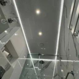 Nowoczesna łazienka z podświetlanym sufitem napinanym, marmurowe płytki, szklana kabina prysznicowa i minimalistyczne oświetlenie. Wykończenie z dbałością o detale.