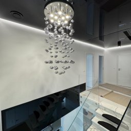 WoodyMAXX - Nowoczesne wnętrze: czarny, lustrzany sufit z oświetleniem LED, kryształowy żyrandol, białe ściany i marmurowe schody z czarnymi nakładkami. Minimalizm i elegancja.