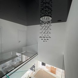 WoodyMAXX Sergiusz Proń - Nowoczesne wnętrze z czarnym, lustrzanym sufitem napinanym i designerskim żyrandolem. Widok z góry na szklane balustrady i częściowo zabezpieczone podłoże.