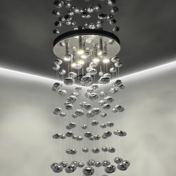 WoodyMAXX Sergiusz Proń - Nowoczesny żyrandol z metalowymi kulami zawieszonymi na cienkich linkach, oświetlający minimalistyczne wnętrze z ukrytym oświetleniem LED.