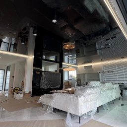 WoodyMAXX Sergiusz Proń - Nowoczesny salon w trakcie wykańczania: czarny, lustrzany sufit z oświetleniem LED, marmurowe podłogi, meble zabezpieczone folią. Widoczne prace wykończeniowe.
