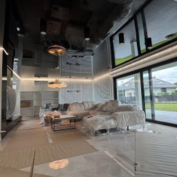 WoodyMAXX Sergiusz Proń - Nowoczesny salon w trakcie wykończenia: marmurowe schody, błyszcząca podłoga, osłonięte meble i duże okna z widokiem na zewnątrz. Oświetlenie LED podkreśla architekturę.