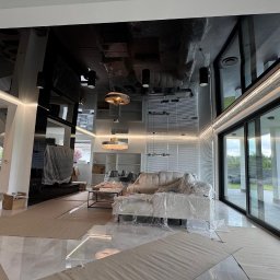 WoodyMAXX Sergiusz Proń - Nowoczesny salon z błyszczącym, czarnym sufitem podwieszanym i dużymi przesuwnymi drzwiami tarasowymi. Meble zabezpieczone folią, widoczne prace wykończeniowe.