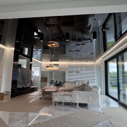WoodyMAXX Sergiusz Proń - Nowoczesny salon po remoncie z błyszczącym, czarnym sufitem napinanym i dużymi oknami przesuwnymi. Meble zabezpieczone folią malarską, podłoga z płytek.