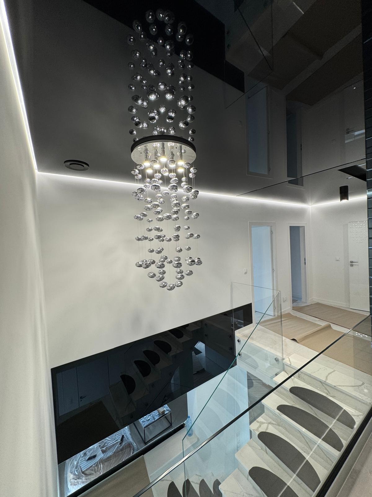 Nowoczesne wnętrze: czarny, lustrzany sufit z designerskim żyrandolem, białe ściany z oświetleniem LED, szklane balustrady schodów i marmurowe stopnie.