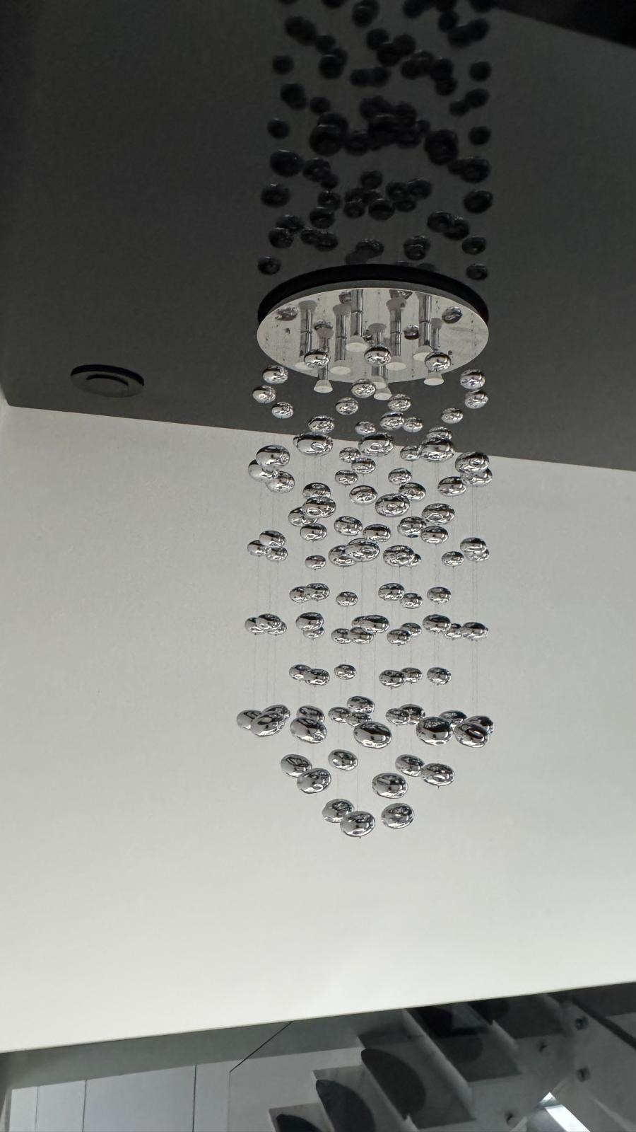 Nowoczesny, chromowany żyrandol 'deszcz' z kulkami zawieszonymi na cienkich linkach, zamontowany na czarnym suficie podwieszanym w jasnym wnętrzu.